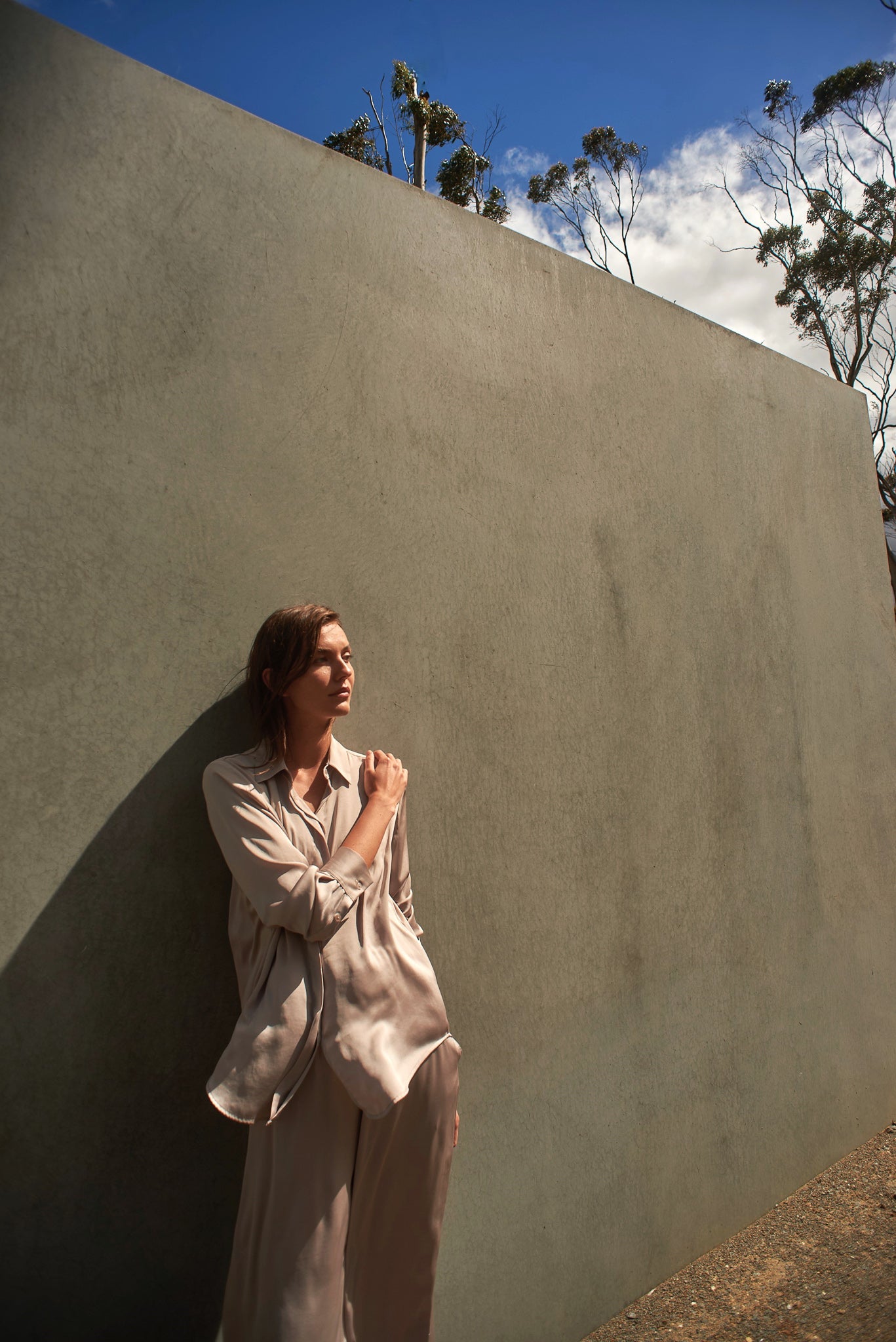 Woman in a beige outfit standing against a textured wall with trees in the background